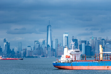 Wall Mural - The ship's stern takes focus as it glides through the bay, with New York City's skyline blurred in the background. The cloudy sky casts a soft, steel-blue tone over the entire scene.