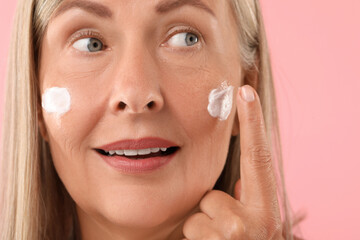 Poster - Senior woman applying face cream on pink background, closeup