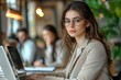 © Pooja - Side view of brunette business woman in glasses wearing beige suit working on laptop while sitting at table in company office with colleagues in, Generative AI
