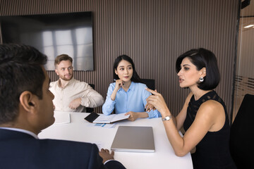 Sticker - Confident attractive team leader woman speaking at meeting with diverse employees, project managers, stakeholders, sitting at large table at boss place. Multiethnic team discussing collaboration