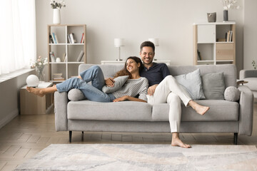 positive happy young husband and wife relaxing on comfortable couch together, talking, hugging, laug