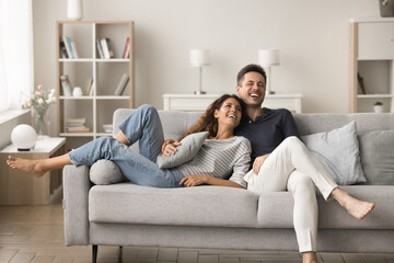 positive happy young husband and wife relaxing on new comfortable pale grey sofa, hugging, talking, 