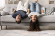 © fizkes - Happy carefree young couple resting upside down on comfortable couch, lying with legs on back and heads hanging to carpeted floor, looking at camera, smiling, laughing, having fun