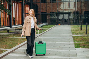 Cheerful young woman in casual outfit walking on the city park with green luggage. 30s Woman dragging suitcase near hotel. Travel Fashion.