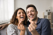 © fizkes - Cheerful happy couple looking at camera, laughing, showing finger heart shapes, symbol of love, happiness, romance. Husband and wife relaxing on home couch together, having fun