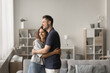 © fizkes - Happy young family couple of homeowners standing in comfortable home interior, hugging in new apartment with love, looking at window away, thinking, planning, talking, laughing