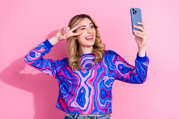 Wall Mural - Selfie photo of cute dyed curly hair girl wearing bright stylish shirt using smartphone cover face v sign pose isolated on pink color background