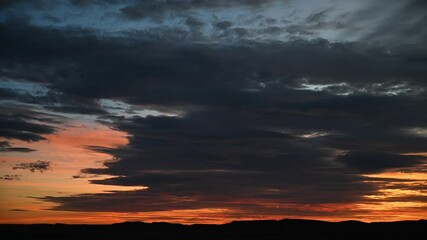 Wall Mural - time lapse of dark clouds in an orange sunset sky with distant silhouetted hills