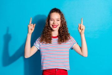 Wall Mural - Joyful woman with curly hair pointing upwards against blue background, expressing positivity and excitement in casual shirt