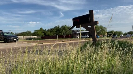 a mailbox on the street of a neighborhood with the grass moving with the wind blowing in a sunny day