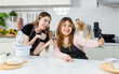 © Bangkok Click Studio - Two happy young beautiful Asian female, woman wearing aprons using mobile phone streaming live video clip or taking selfie photo smiling while making bakery in kitchen enjoy teenage lifestyle at home.
