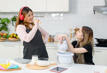 Two happy young beautiful Asian female, woman wearing aprons relax listen headset music looking tablet recipe guidebook smiling while making bakery in kitchen enjoy teenage lifestyle at home