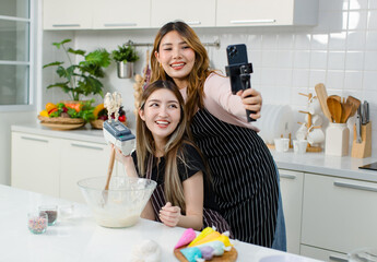 Two happy young beautiful Asian female, woman wearing aprons using mobile phone streaming live video clip or taking selfie photo smiling while making bakery in kitchen enjoy teenage lifestyle at home.