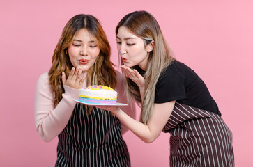 Photo two cute happy fun young Asian beautiful female, woman smiling wearing aprons holding cakes sweet dessert poses isolated plain pastel light over pink color wall background.