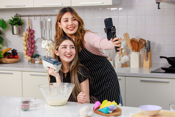 Two happy young beautiful Asian female, woman wearing aprons using mobile phone streaming live video clip or taking selfie photo smiling while making bakery in kitchen enjoy teenage lifestyle at home.