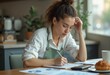© kotlyarn - A woman in an apron reviews financial documents while sitting at a table in a bright cafe.