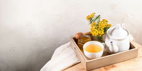 Wall Mural - Tansy tea in teapot and cup, tansy flowers, honey in wooden tray on textured white wall backdrop.