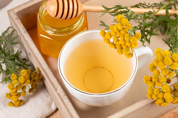 Wall Mural - White cup with healthy medicine tansy tea, honey and tansy flowers in wooden tray close up.