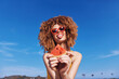 © SHOTPRIME STUDIO - Cheerful young woman holding a slice of watermelon under a sunny blue sky