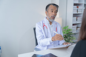 Senior male doctor is explaining a diagnosis to a patient while holding a clipboard and gesturing with a pen in his office