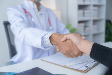 Doctor and patient shake hands in an office, showing trust and forming a healthcare partnership. The doctor provides care and advice, with paperwork and a clipboard in the background