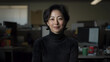 © Nattapol - woman with short black hair wearing black turtleneck sweater smiles confidently in office environment, surrounded by computer screens and green cup