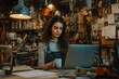 © Design Dynasty - Woman working in workshop holding mug while looking at laptop for design work surrounded by various tools and materials, Generative AI