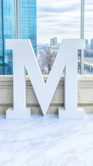 A large white letter M sits on a marble floor in front of a window with a city view.