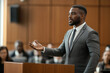 © tynza - Confident African-American male lawyer in a modern courtroom, presenting arguments in front of a jury.