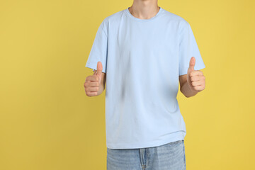 Sticker - Teenage boy wearing light blue t-shirt and showing thumbs up on yellow background, closeup. Space for text