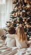 © Maria Mikhaylichenko - Cozy Christmas morning scene: two children in white sweaters sit on a bed, gazing at a beautifully decorated tree with golden ornaments and twinkling lights.