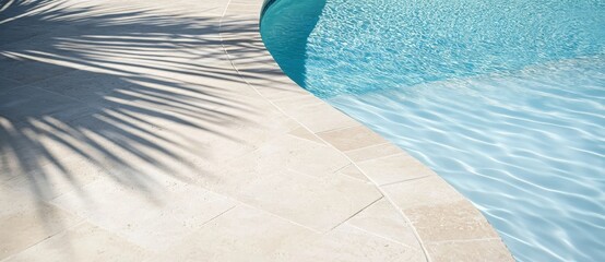 Close-up of light beige travertine tiles around the edge of an outdoor pool, with a palm tree's shadow on one side and clear blue water in the background. 