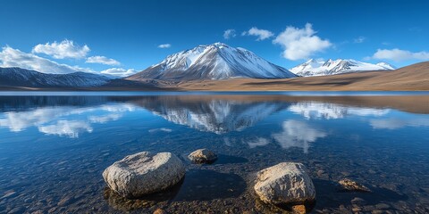 Clear blue mountain lake reflecting snow-capped peaks and fluffy clouds in a tranquil scene with smooth water and large rocks.