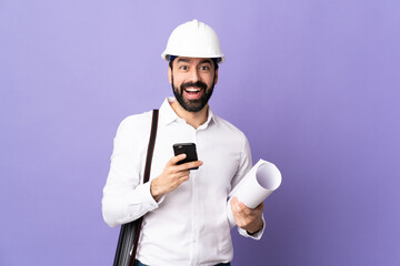 Wall Mural - Young architect man with helmet and holding blueprints over isolated purple background surprised and sending a message