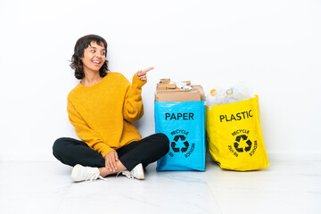 Wall Mural - Young girl holding a bag full of plastic and paper sitting on the floor isolated on white background pointing finger to the side and presenting a product