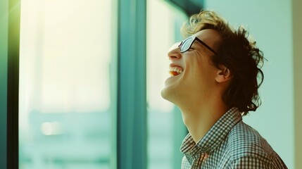 Sticker - A joyful young man wearing glasses laughs heartily by a window, bathed in warm sunlight.