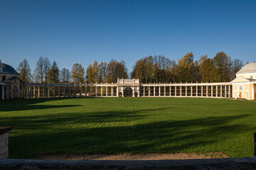 The image shows a neoclassical colonnade under restoration, with scaffolding surrounding the white columns. Trees with autumn leaves and a clear blue sky form the natural backdrop.