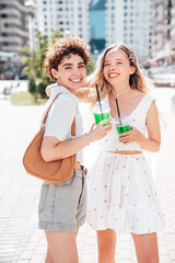 Two young beautiful smiling hipster female in trendy summer clothes. Carefree women posing outdoors. Positive models holding and drinking fresh cocktail smoothie drink in plastic cup