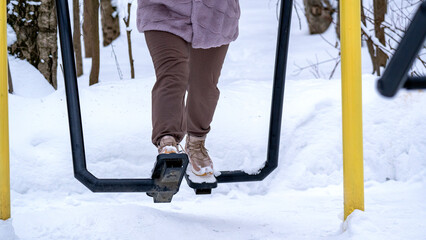 Woman in the park in winter on the exercise machine. Healthy smiling woman exercise on outdoor body builder trainer machines in winter time. Snow falling in park