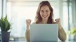 © BoOm - Happy young woman celebrating success in front of her laptop in a bright office environment.