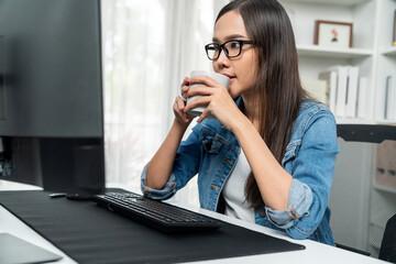 Working young asian creative manager with glasses focusing on pc while drinking hot coffee, creating with website design on social media online with blue jeans shirt at home office. Stratagem.