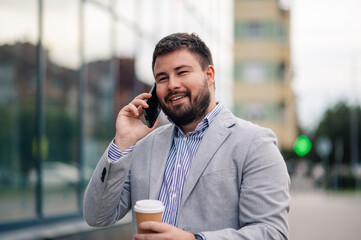 Wall Mural - Businessman smiling while talking on the phone and holding coffee outside office building