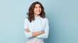 © Iona - Young woman with curly hair in blue blouse stands confidently, arms crossed, against a neutral backdrop, radiating self-assured happiness.