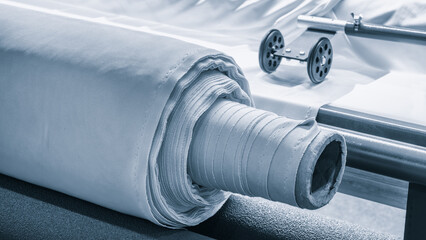 Poster - A roll of fabric is laid out on a table next to a sewing machine. The machine is likely used to cut and stitch the fabric into a finished product. The roll of fabric is unrolled and he is quite large