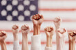 © barmaleeva - Hands with raised fists show solidarity and unity in front of a blurred American flag, symbolizing strength, activism, and diversity.