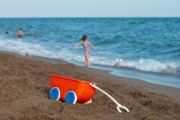 Red colored toy on sand on beach. Girl playing in the background and blue sea.