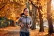 © miss irine - Young woman jogging in autumn park. Wears gray jacket, blue pants, smiling running. Park around displays vibrant autumn colors with orange, yellow leaves. Woman path leads viewer eye towards right