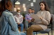 © miss irine - Group therapy session with female psychologist and clients. Adult people discuss mental health issues in indoor office setting. Pro woman leads discussion with clipboard, notes on whiteboard.