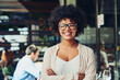 © SometimesNever/peopleimages.com - Arms crossed, portrait and smile of business black woman outdoor at coffee shop for career development. Design, face and glasses with happy employee at cafe or restaurant for creative startup