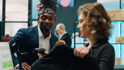 Poster - African american shopper buying an elegant suit jacket from clothing store, choosing a formal fashion item to create an outfit. Retail employee at cash register scanning product to sell. Camera A.